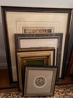 Stack of six framed artworks in various frame styles leaning against a wall