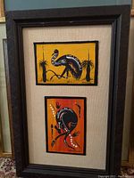Full view of the two framed artworks together, showing the bird and frog paintings in same frame with beige matting and textured dark frame.