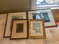 Four framed artworks arranged on a tufted bench near a window: a watercolour floral painting, two animal prints, and a signed porch scene print.