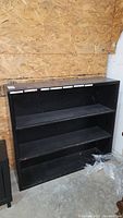 Front view of black wooden bookcase showing three shelves and overall condition against plywood background