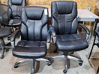 Two black adjustable rolling desk chairs side by side in a warehouse setting.