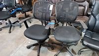 Two black mesh-backed office chairs with padded seats side by side among other chairs in a warehouse.
