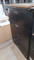 Side and front angled view of the black HON metal filing cabinet with four drawers, showing surface scratches and wear.