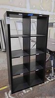 Front view of black laminate shelving unit with 8 cube compartments, showing some scratches and overall used condition.