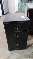 Front view of the black laminate storage cabinet showing three drawers with metal handles and label holders.