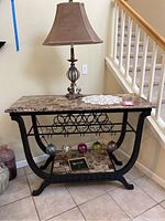 Full view of the bar table with polished stone top, black metal frame, lamp on top, and visible glass stemware ornaments hanging from the rack below the tabletop.