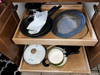 Drawer with two black pans and several lids including clear glass lids with metal rims, and one white saucepan with black handle.