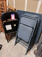 Stack of four gray padded folding chairs with metal frame next to the folding table and one black padded folding chair visible