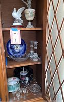 Lower shelf with six small clear stemmed glasses, a decorative ceramic plate with floral design, and black stemware with handle.