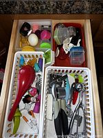 Open drawer with more kitchen utensils including plastic spoons, bottle openers, garlic press, scoops, and small kitchen tools, plus several small plastic containers with lids.