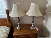 Two matching silver-toned table lamps with fabric shades on wooden nightstand next to bed