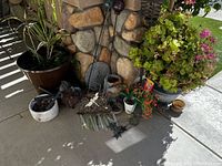 Photo of assorted potted plants and garden decor including flowers, greenery, lantern, and garden stakes placed by stone wall on outdoor porch area.