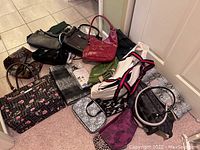 Overview photo of multiple handbags and purses lying on carpeted floor showing variety of colors and styles.