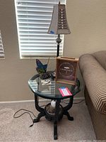 Round glass top side table with butterfly lamp, wine cork collector, and lamp cord visible on carpeted floor near window and sofa.