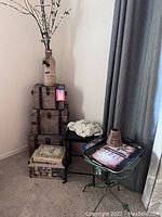 Photo showing stacked decorative storage boxes, a tall faux flower vase with white blossoms, a dark wood square table, and a glass top metal table.