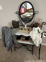 Frontal view of vanity table with mirror, lace cloth, perfume bottles, bench, and assorted clothing items draped over.