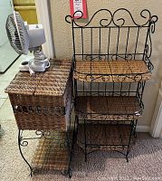 Three-tier wicker shelving unit next to wicker side table with white oscillating fan on top.