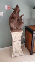 Seated ceramic figure playing a lute, brown-toned, sitting on a cream-colored ornate pedestal with lattice designs, size around 27 inches tall for the figure and 34 inches tall for the pedestal.