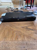 Front angle of Sony Blu-Ray DVD player on wooden surface showing brand and model detail with cables.