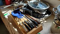 Photo showing Calphalon stainless steel saucepans and skillet with glass lids stacked, along with blue enameled spoon rest and knife block with cutlery on countertop