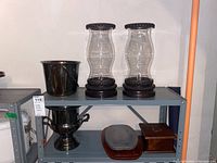 Two glass and metal candle holders with clear curved glass and ornate black metal bases and tops placed on a shelf alongside silver plated buckets and a wooden box.