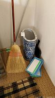Tall blue and white ceramic vase with intricate floral designs placed on carpet near wall, alongside broom handle and dustpan handle visible behind it. Two containers of disposable cleaning sheets nearby.
