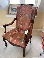 Front left angled photo of the upholstered chair showing wooden carved arms, legs, and patterned fabric upholstery with vintage style figures.