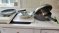 Three pans including a non-stick frying pan, skillet with glass lid, and deep sauté pan with lid are arranged side by side on a white kitchen counter.