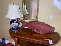 Full view of wooden fish sculpture and glass base lamp side by side on wooden table beneath mirror.