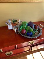 Lot contents on wooden furniture with glass vegetables on silver platter and cut-glass bowl with additional glass vegetables inside.