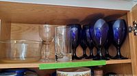 Wood cabinet shelf with 8 cobalt blue goblets lined up behind a beer glass mug and a clear glass bowl.