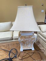 Full view of the white table lamp with gold tone Japanese decorative motifs on the base and cream fabric shade, showing missing finial on top.