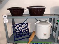 Two brown ceramic Dutch ovens with lids on top shelf; lower shelf holds blue metal book stand, wooden rolling pin, and white Aroma rice cooker.
