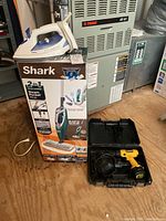 Photo showing Shark Steam Mop box, T-Fal iron on top, and Dewalt drill in black hard case on wooden floor
