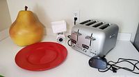 Photo showing all items: DeLonghi 4-slice toaster, Taylor scale behind, black digital alarm clock, large ceramic pear, and red ceramic platter.