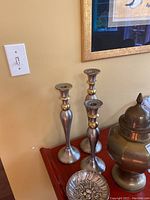 Three silver tone candlesticks with brass accents and a silver tone decorative dish on a wooden table.