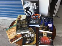 Photo showing seven hardcover books placed on a round table, visible titles include Moscow Rules, Last Snow, The Silkworm, and more.