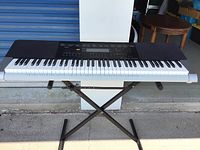 Full view showing Casio WK-240 keyboard placed on a black metal X-style adjustable stand outside near a blue storage door.