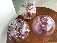 Three decorative glass globes on wooden surface, two clear with internal red and white floral designs, one solid light purple.