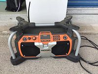 Front view of the RIDGID orange and black jobsite radio with protective corners and handle, placed on concrete surface.