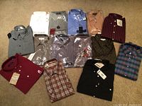 Overview of all 14 men's shirts arranged on carpet. Mixture of new in package and folded shirts in various patterns and colors.