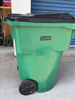 Photo of large green utility bin with black hinged lid and two wheels, shows ULINE logo on front and visible scratches.