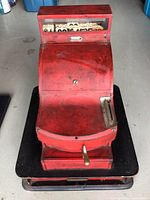 Front top angled view of the vintage red metal cash register, showing the curved top, brass lever, and black drawer.