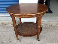 Full view of the wooden table showing round top, carved apron, four legs with caster wheels, and oval lower cane shelf.