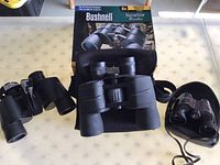 Three pairs of binoculars displayed on a table: Bushnell NatureView with box and case in center, Taylor on left, Nikon on right with strap and case.