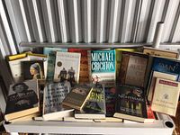 Photo showing an array of used hardcover books, some stacked and others standing, including recognizable titles by Leonard Cohen, Dan Brown, Michael Crichton, Stieg Larsson, and others.