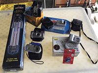 Overview shot of tripod box, Brownie Hawkeye camera and box, Nikon Coolpix box, assorted compact cameras and camera cases, SanDisk memory card on table.