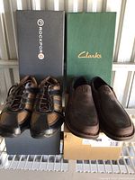 Two pairs of men's shoes side by side with their original boxes behind. Left pair is Rockport multi-tone sneakers, right pair is Clarks brown slip-ons.