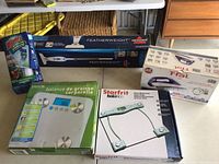 Multiple houseware items displayed together including Starfrit glass scale, Taylor body fat scale, Bissell Featherweight vacuum, T-Fal iron, and Windex glass cleaner spray.