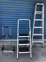 Photo of two ladders and a folding dolly against blue wall. Includes a white three-step ladder with black treads, a taller aluminum ladder, and a folded dolly.